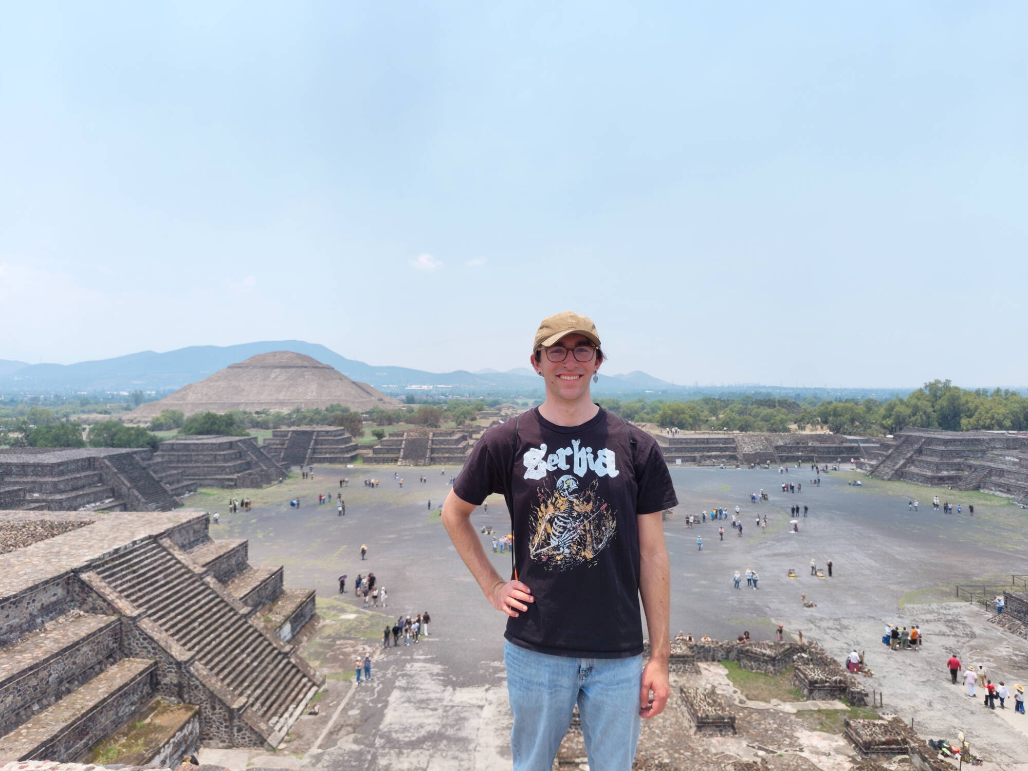 Andrew is wearing blue jeans and a "Serbia" black t-shirt with a light brown baseball cap. They are smiling with dark rimmed glasses